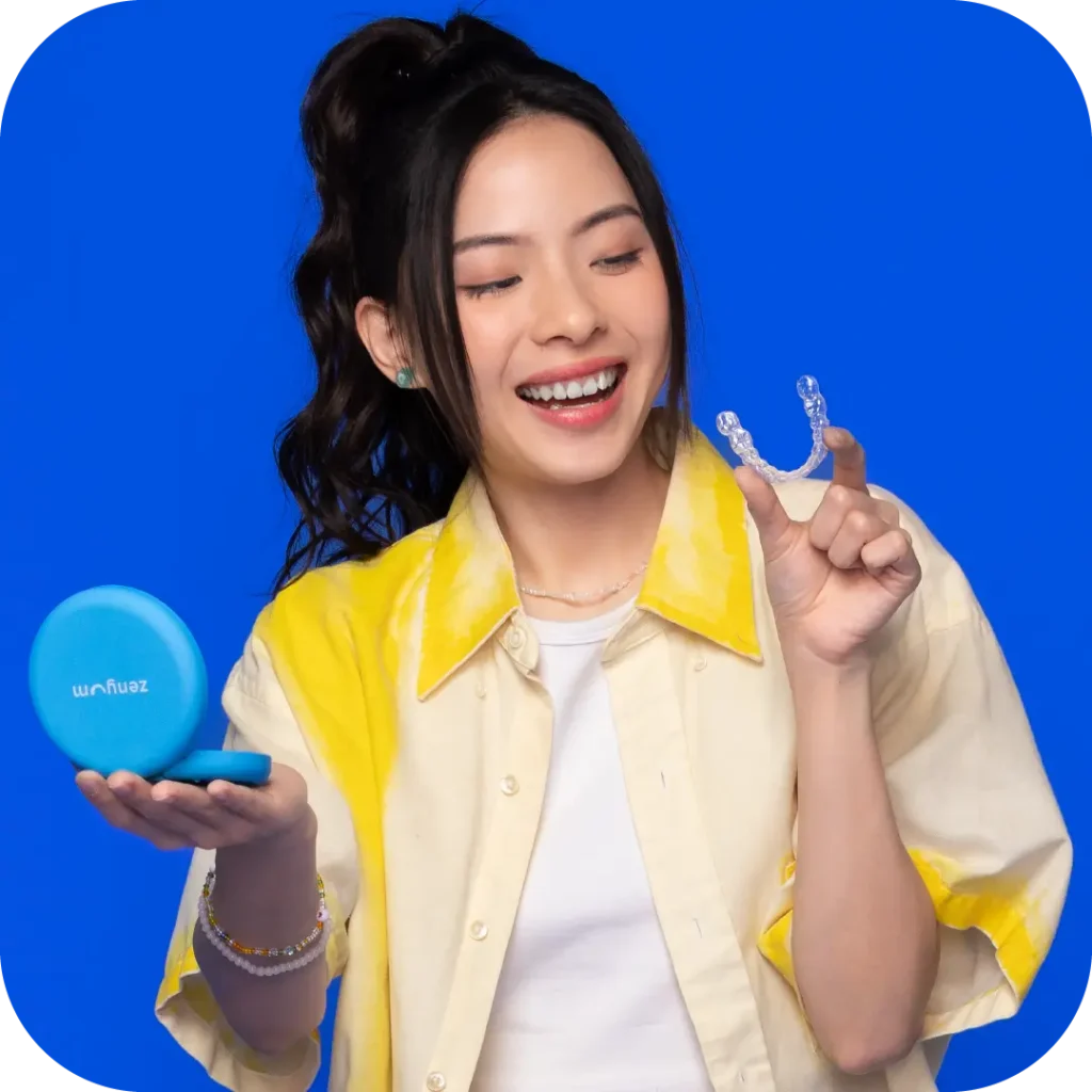 A girl wearing a yellow top holding Zenyum Invisible Braces while smiling against a blue backdrop.