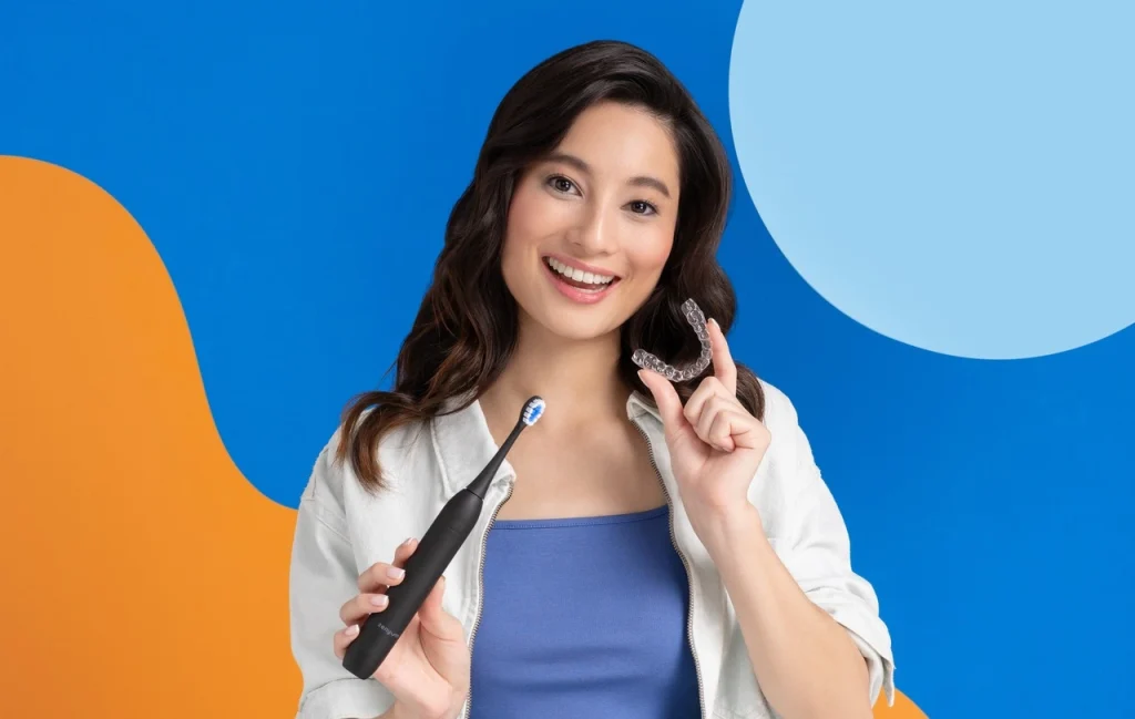 Person holding a ZenyumSonic™ electric toothbrush and Zenyum Invisible Braces, against a vibrant orange and blue backdrop.